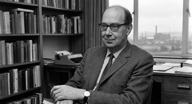Middle-aged man wearing glasses and suit sitting in a study with bookshelves, typewriter, and paperwork